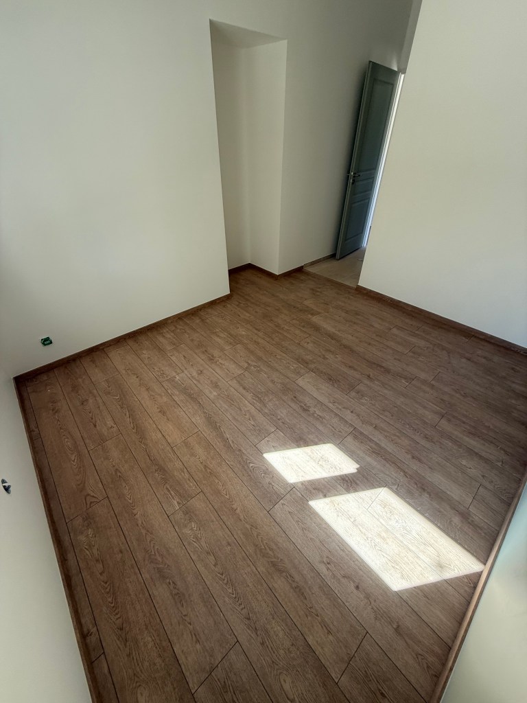A spacious empty room with wooden flooring, featuring a doorway and natural light streaming through a window.